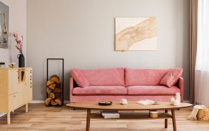 Sala de estar pequena composta por paredes cinza, sofá rosa, mesa de centro de madeira, lenhas empilhadas, balcão, cesta de toalhas e quadro abstrato na parede, utilizando as melhores cores para aumentar a amplitude do espaço.